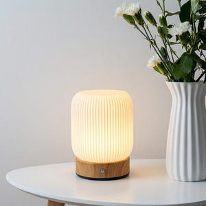 Może przedstawiać: Nowoczesna, cylindryczna lampa stołowa z żebrowanym, kremowym kloszem i drewnianą podstawą. Lampa jest podświetlona, emitując ciepłą poświatę. Biały wazon z białymi kwiatami stoi obok.