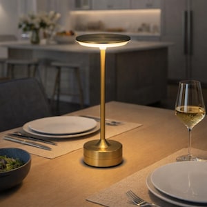 Puede incluir: Una lámpara de mesa dorada con una pantalla circular y un tallo delgado. La lámpara está sobre una mesa de madera puesta para una comida, con platos, cubiertos y una copa de vino. El diseño de la lámpara es moderno y minimalista.