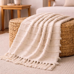 Può includere: Una coperta color crema con un motivo a righe orizzontali testurizzate e bordi con frange annodate, drappeggiata su un pouf rotondo intrecciato in fibre naturali. La coperta presenta una morbida struttura in tessuto con dettagli a frangia.