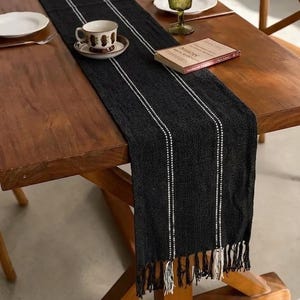 May include: A black table runner with white vertical stripes and a fringed edge. A teacup and saucer, a book, and a plate are on the wooden table. The runner is made of a woven material.