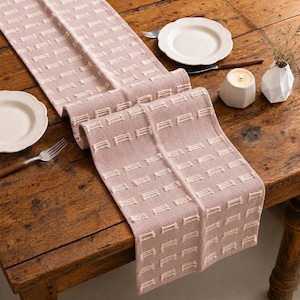 May include: A dusty rose table runner featuring a repeating pattern of small, raised cream-colored rectangular tufts. The fabric has a textured, woven appearance and is draped across a wooden surface, accompanied by white plates and a candle.