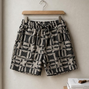 Peut inclure: Un short à motif géométrique répétitif gris anthracite et beige. Le short est doté d'une taille élastique avec un cordon de serrage en tissu assorti et de deux poches plaquées à l'avant, suspendu à un cintre en bois.