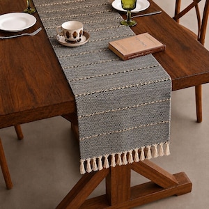 May include: A rectangular charcoal gray table runner with a woven texture, featuring horizontal tan stripes and cream-colored fringe tassels at the ends. The runner is laid flat across a wooden table, providing a base for a cup and a book.