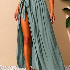 May include: A sage green maxi skirt featuring a high-waisted design with a side tie closure and a long, dramatic leg slit. The fabric has a subtle, crinkled texture, creating a flowing silhouette that extends to the floor in a solid, muted tone.