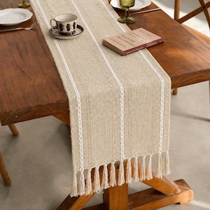 May include: Beige table runner with white vertical stripes and fringe. The runner is made of a textured fabric and is placed on a wooden table. A cup and saucer, a book, and plates are on the table.