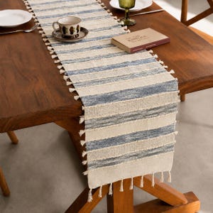 May include: A rectangular table runner featuring a woven pattern of alternating cream and blue horizontal stripes. The runner is finished with small, decorative tassels along both long edges and the bottom edge, creating a textured appearance.