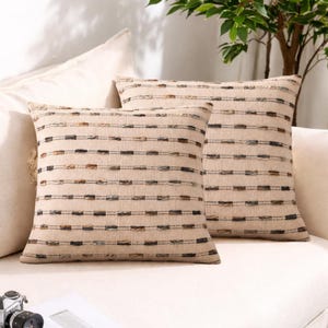 May include: Two beige square throw pillows with horizontal stripes of various colors. The pillows are on a white sofa, with a plant in the background. The pillows have a textured appearance and are likely made of woven fabric.