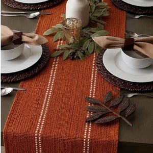 May include: An orange woven table runner with white vertical stripes, set with white plates, bowls, and napkins. Brown woven placemats and napkin rings complement the warm colour scheme. Decorative elements include a leaf-shaped accent and a gold-toned glass.