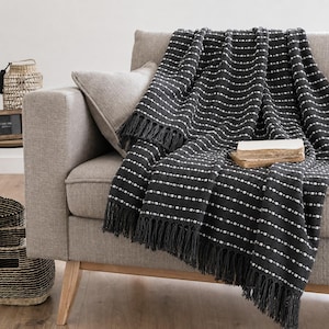 May include: A dark charcoal grey throw blanket featuring horizontal white dotted stripes and long fringe trim along the edges. The blanket is draped over a light beige sofa, with a small, rectangular, light-coloured book resting on top of the fabric.
