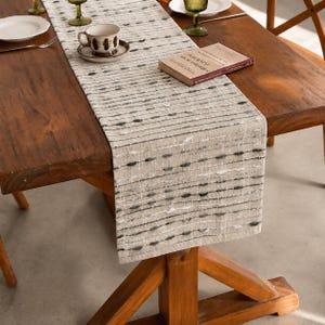 May include: A beige table runner featuring a textured pattern of horizontal dark grey and white dashed lines. The runner is laid across a wooden table, accompanied by a ceramic cup on a saucer and a closed book with visible text on the cover.