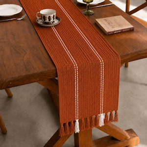 Puede incluir: Un camino de mesa de color óxido con dos líneas verticales punteadas blancas y borlas blancas en los extremos. Se coloca sobre una mesa de madera oscura, con una taza y un platillo visibles.