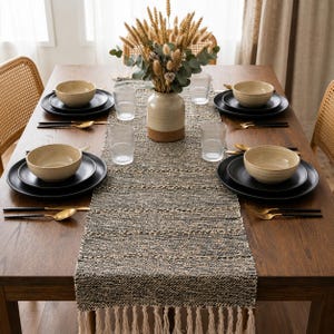 May include: A wooden dining table set with four place settings, each featuring a matte black plate and a beige bowl. A textured black and beige table runner with fringe ends lies down the centre, holding a ceramic vase filled with dried wheat.