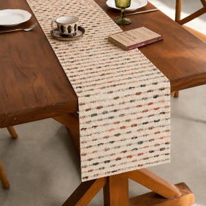 May include: A beige table runner with a textured, woven design featuring small, colorful knots in shades of brown, orange, and gray. The runner is placed on a dark wooden table, with a teacup, saucer, and book visible.