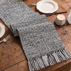 May include: A rectangular table runner featuring a woven texture in a blend of black, white, and gray threads. The runner has a thick, chunky weave pattern and is finished with long, straight fringe tassels along the short edges of the fabric.