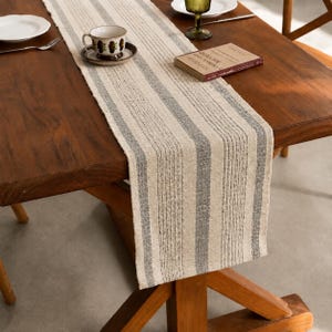 May include: A rectangular table runner featuring a woven texture with alternating vertical stripes in natural beige and dark gray. The fabric has a visible coarse weave pattern, extending across the length of the wooden table surface.