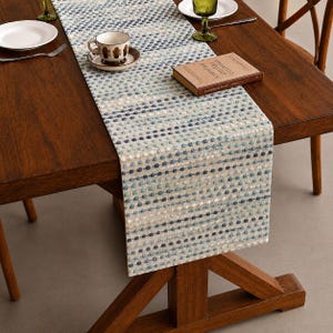 May include: A rectangular table runner featuring a textured, woven pattern of horizontal stripes composed of small, raised dots in varying shades of blue, teal, and white, laid flat across a dark brown wooden dining table surface.