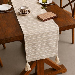 Peut inclure: Un chemin de table rectangulaire présentant un motif à rayures horizontales beige et blanc en tissu tissé. Le chemin de table est agrémenté de pompons décoratifs blanc cassé fixés à chacun des deux coins inférieurs, ajoutant de la texture.