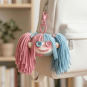 Puede incluir: Un llavero de muñeca hecho a mano con cabello de hilo rosa y azul, ojos de botón y boca cosida. La muñeca está sujeta a un clip de llavero rosa, colgando de una mochila blanca. La cara de la muñeca es de tela.
