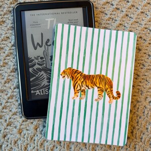 Peut inclure: Un lecteur d'ebook noir affichant le texte de "The We" d'Alistair, à côté d'un carnet à rayures vertes et blanches avec une illustration de tigre. L'ensemble est posé sur une surface texturée.