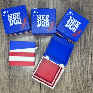 Puede incluir: Tres cajas cuadradas azules con el texto blanco NEE DOH y gráficos de estrellas rojas en las tapas, una caja cerrada con rayas rojas y blancas en el lateral y una caja abierta con tapa azul e interior rojo, todas con un esquema de colores patriótico.