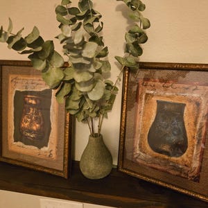 May include: Two framed art prints with bronze-colored frames, each featuring a vase illustration. A small, green vase holds eucalyptus branches. The artwork is displayed on a dark brown shelf against a cream-colored wall.