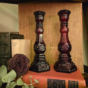 May include: Two ornate, dark red glass candlesticks with detailed patterns. They stand on a red book with gold lettering, next to a dried rose and old books. The candlesticks have a vintage, antique aesthetic.