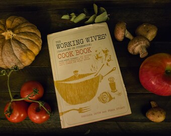 Może przedstawiać: Książka kucharska w stylu vintage zatytułowana The Working Wives' Cook Book z kremową okładką, na której znajduje się żółta ilustracja garnka, widelca i solniczki, otoczona dynią, czerwonymi pomidorami, grzybami i czerwonym jabłkiem.