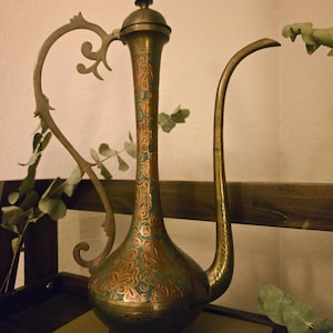 May include: A tall brass pitcher with a long, slender neck and a curved spout. The metal surface features intricate floral engravings highlighted with teal-colored enamel accents. It has a decorative, ornate handle and a matching lid with a finial.