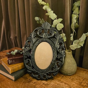 May include: An oval picture frame with a glossy black finish and intricate, symmetrical carved leaf-like patterns surrounding the central opening. The frame features a decorative top crest and a smooth, light tan backing visible within the oval center.