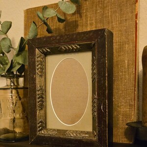 May include: A rectangular dark brown wooden picture frame with a distressed finish and an oval-shaped inner matting. The frame features carved geometric chevron patterns along the inner border, surrounding a plain, light-colored oval insert.