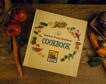 Może przedstawiać: Książka kucharska The General Foods Kitchens COOKBOOK to kwadratowa pozycja w twardej oprawie o kremowym kolorze, z okrągłym układem kolorowych ilustracji żywności oraz centralnym logo z łyżką, filiżanką i napisem General Foods Kitchens.
