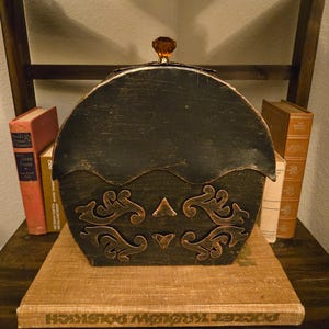 May include: A black and gold decorative box with an ornate design, topped with a brown gem. The box is sitting on a wooden shelf with books in the background. The box has a curved top and a decorative front.