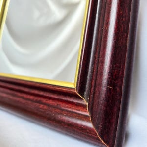 Może przedstawiać: Prostokątne lustro z drewnianą ramą w kolorze głębokiego mahoniu o gładkim, polerowanym wykończeniu. Wewnętrzna krawędź ramy jest podkreślona cienką, złotą metalową obw&oacute;dką, kt&oacute;ra otacza odblaskową szklaną powierzchnię lustra.