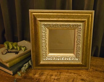 May include: A square mirror featuring a wide, distressed gold-toned frame with a secondary inner border displaying a raised, repeating scrollwork pattern in a lighter, metallic silver-gold finish, surrounding a clear, reflective glass center.