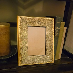 May include: A rectangular picture frame with a textured, mottled surface in shades of green, gold, and brown. The frame features a smooth, thin gold border along the inner and outer edges, surrounding a plain, light-colored rectangular insert.