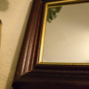 Może przedstawiać: Prostokątne lustro w ciemnobrązowej drewnianej ramie o gładkim, zaokrąglonym profilu. Wewnętrzna krawędź ramy jest podkreślona cienką, złotą metalową listwą, kt&oacute;ra obramowuje powierzchnię lustra, tworząc wyraźny kontrast między elementami.