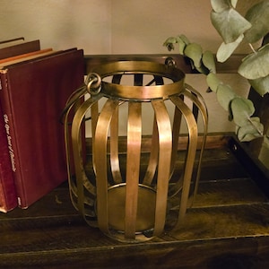 May include: A cylindrical lantern constructed from vertical brass-toned metal slats. The lantern features a circular base, an open top, and a small metal ring attached to the upper rim for hanging. The metal has a smooth, reflective finish.