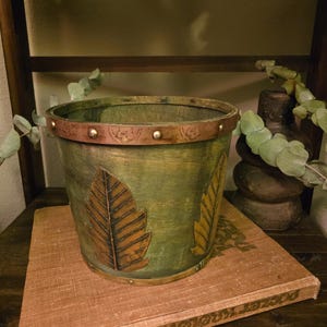 May include: A cylindrical green planter featuring a copper-toned metal rim with small studs and embossed leaf motifs. The body of the planter has a brushed finish, and the exterior is decorated with two prominent, raised copper-colored leaf appliques.