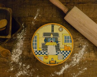 Może przedstawiać: Okrągła drewniana pokrywka z kolorową ilustracją osoby w kuchni trzymającej znak z napisem COOKIE CUTTERS, z kotem, zegarem ściennym i szachownicą na podłodze, z napisem LILLIAN VERNON.