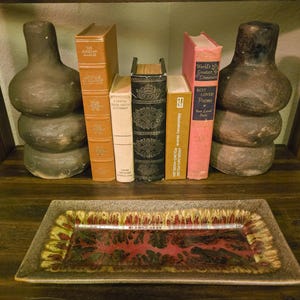 May include: A wooden shelf displays decorative items. Two stacked, bulbous, gray-brown vases flank a row of vintage books in various colors. A rectangular ceramic tray with a red and yellow pattern sits on the shelf below.