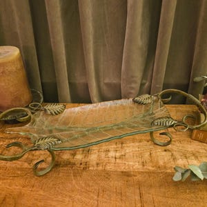 May include: A rectangular glass tray with a leaf-vein texture, held by a dark bronze-toned metal frame featuring decorative scrollwork and metal leaf accents at the corners. The tray is elevated on four curved metal feet and has two handles.