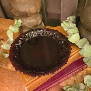 May include: A round, dark purple glass plate featuring a scalloped edge and a decorative border with a repeating oval and cross-hatched pattern. The center of the plate displays a subtle, radiating sunburst design embossed into the glass surface.