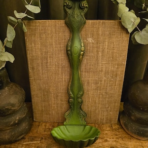May include: A tall, vertical wall-mounted basin or font in a matte olive green finish. The piece features an elongated, ornate backplate with embossed scrollwork and a shield-like emblem at the top, leading down to a scalloped, shell-shaped bowl.