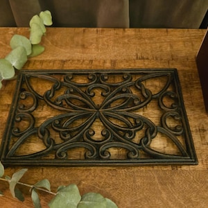 May include: A rectangular cast iron trivet featuring a dark brown finish and an intricate, symmetrical openwork scroll pattern. The metal piece has a flat, grid-like structure with curved, interconnected lines forming a decorative design.