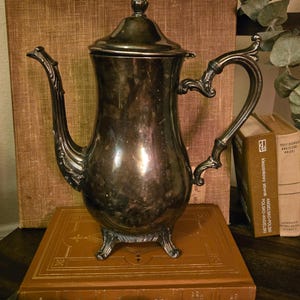 May include: An antique silver-colored coffee pot with ornate detailing, including a curved handle and spout. The pot sits atop a vintage book with gold lettering, with other books and foliage in the background. A classic piece for home decor.