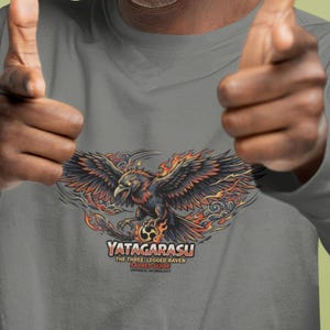 Pode incluir: Uma camisa cinza de manga comprida com um gráfico detalhado de um corvo de três pernas com chamas e o texto "YATAGARASU THE THREE LEGGED RAVEN SACRED GUIDE". A pessoa está usando um gorro cinza e óculos.