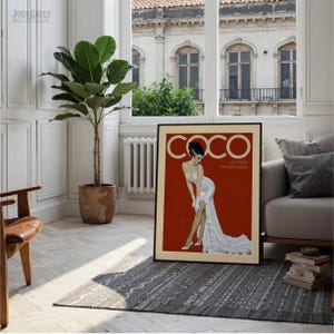 a large framed poster of a woman in a white dress, with the word "COCO" prominently displayed. The poster is placed on a rug in a living room setting, with a potted plant and a window visible in the background.
