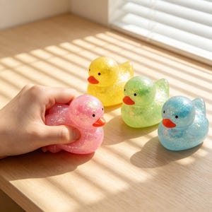 Könnte beinhalten: Vier glitzernde Entenspielzeuge in Gelb, Grün, Blau und Rosa. Die rosa Ente wird von einer Hand gehalten. Jede Ente hat einen roten Schnabel und schwarze Augen. Die Enten sitzen auf einer hellbraunen Oberfläche.