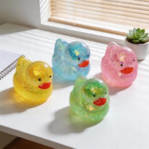 Peut inclure: Quatre canards en plastique remplis de paillettes jaunes, bleus, roses et verts. Chaque canard a un bec rouge et de petits yeux noirs. Les canards sont posés sur une surface blanche près d'une fenêtre.