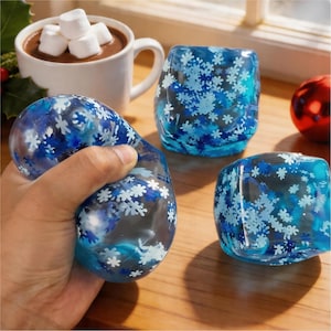 May include: A hand squeezing a clear, blue stress ball filled with blue and white snowflake shapes. Two other blue snowflake-patterned objects are on a wooden surface, along with a mug of hot chocolate topped with marshmallows.